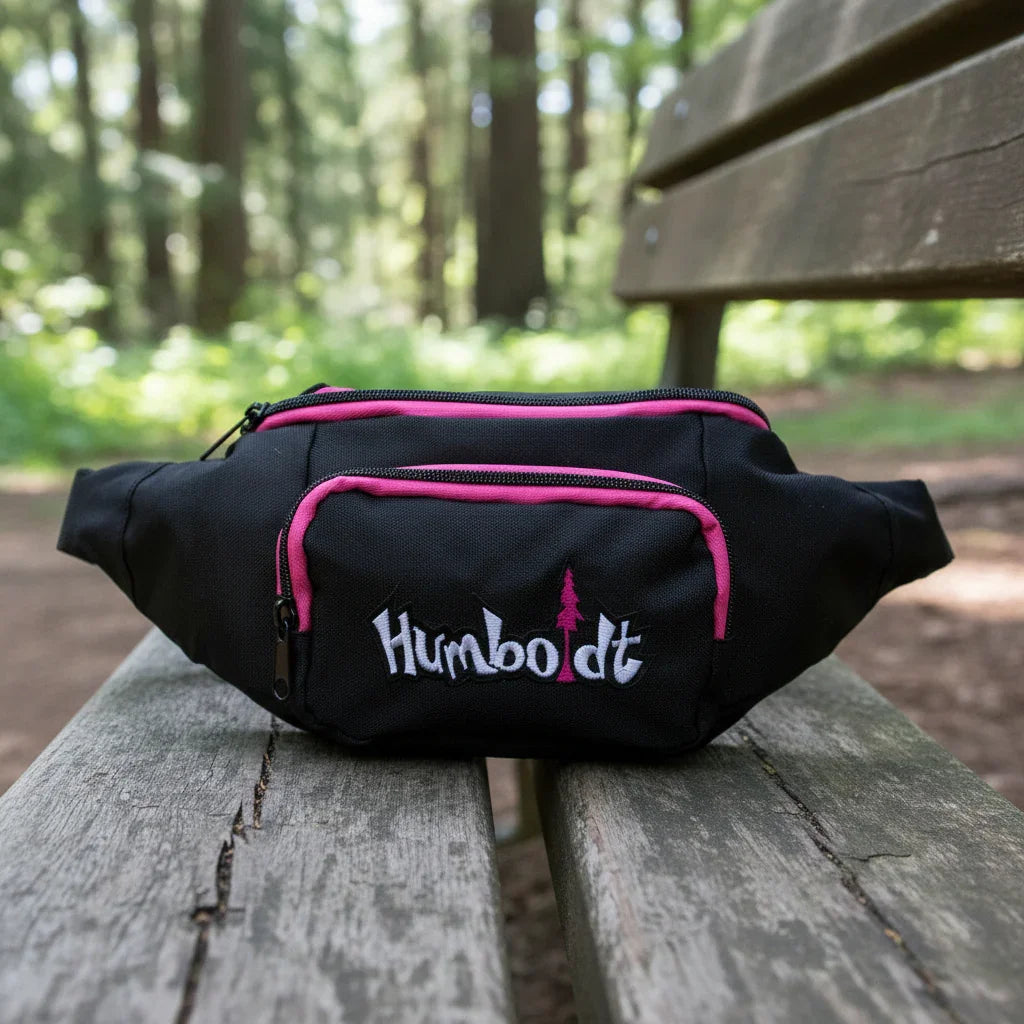 Black fanny pack with pink zippers and Humboldt logo, resting on wooden bench in forest setting