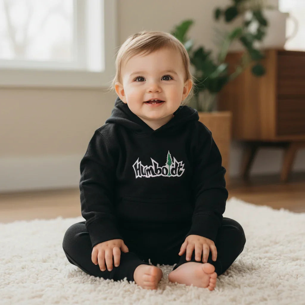 Baby wearing black Humboldt hoodie with tree logo, sitting on white rug in cozy room